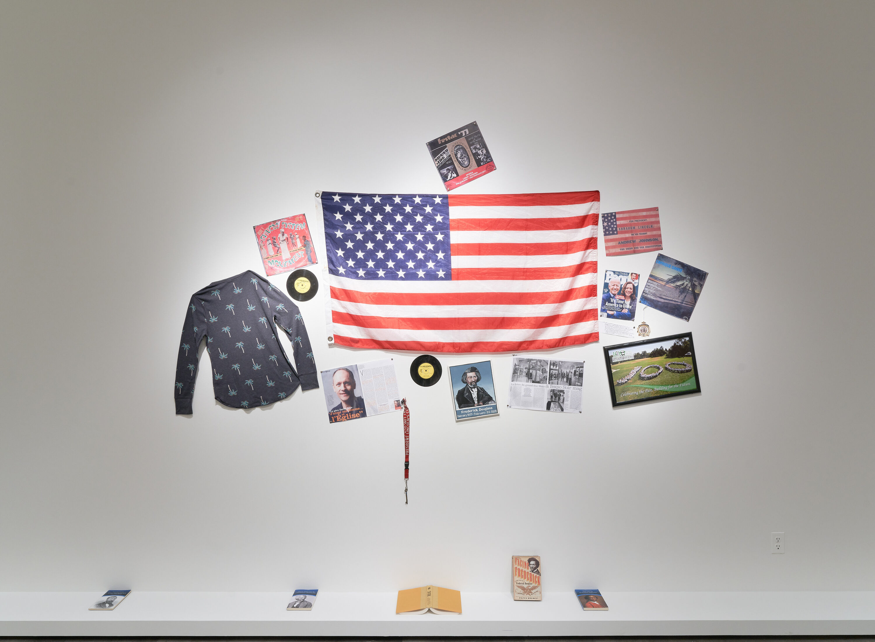 on a white wall an american flag, portrait of Frederick Douglass and books on a ledge along the floor