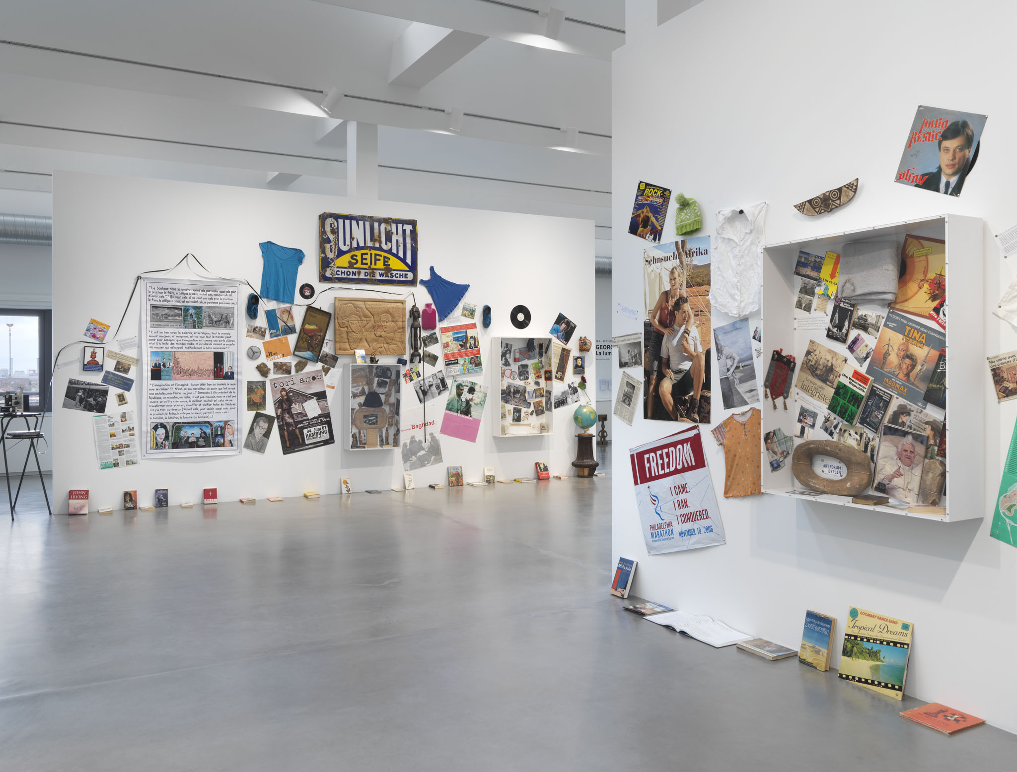 two walls in a large space with dozens of objects: used textiles, posters, hand-painted banners, books on the floor around the ledges