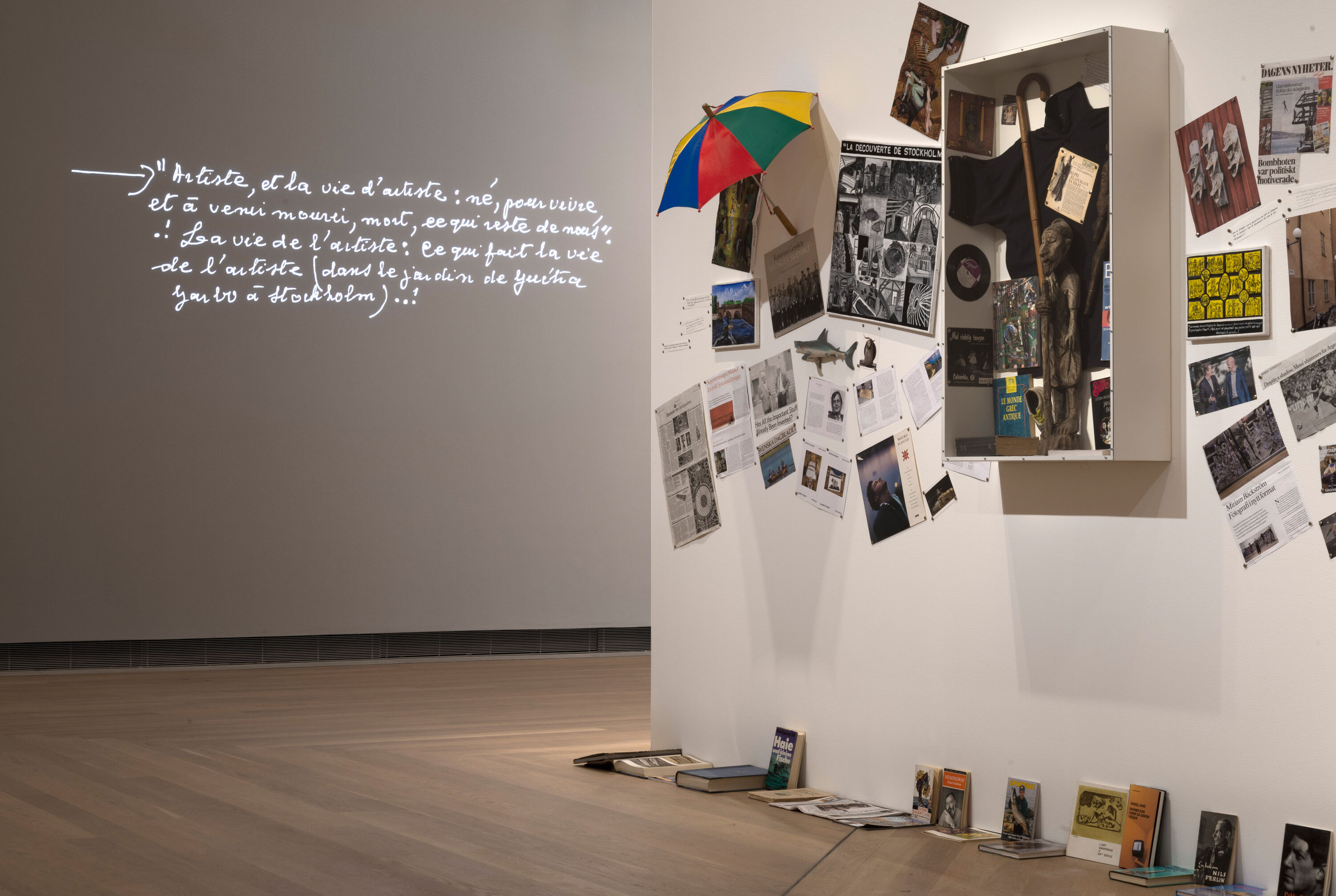 installation of paintings, books and umbrella on white wall. Handwritten Text projected on the wall.