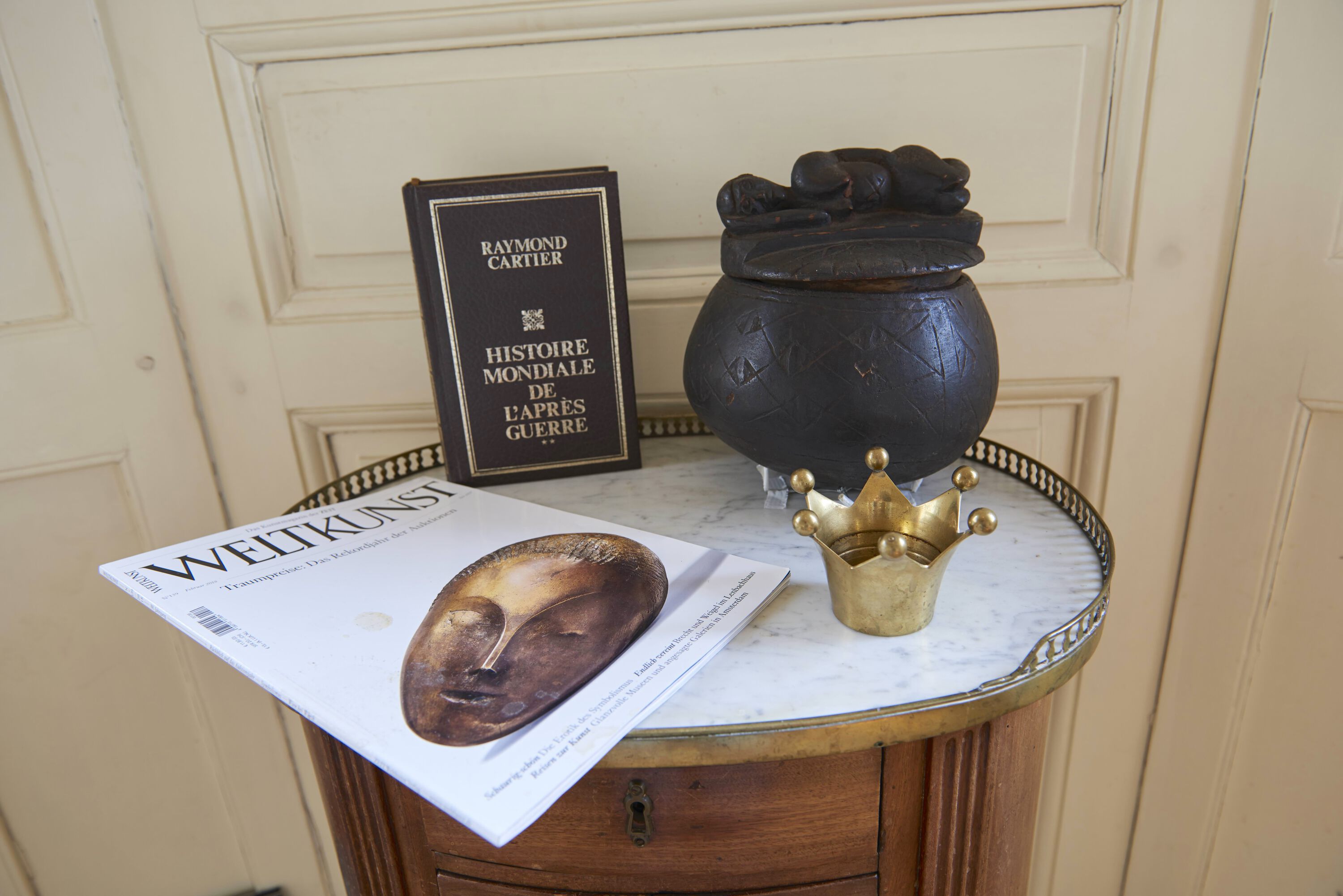 on a marble table an art magazin, a book, a brass crown and an african tobacco pot.