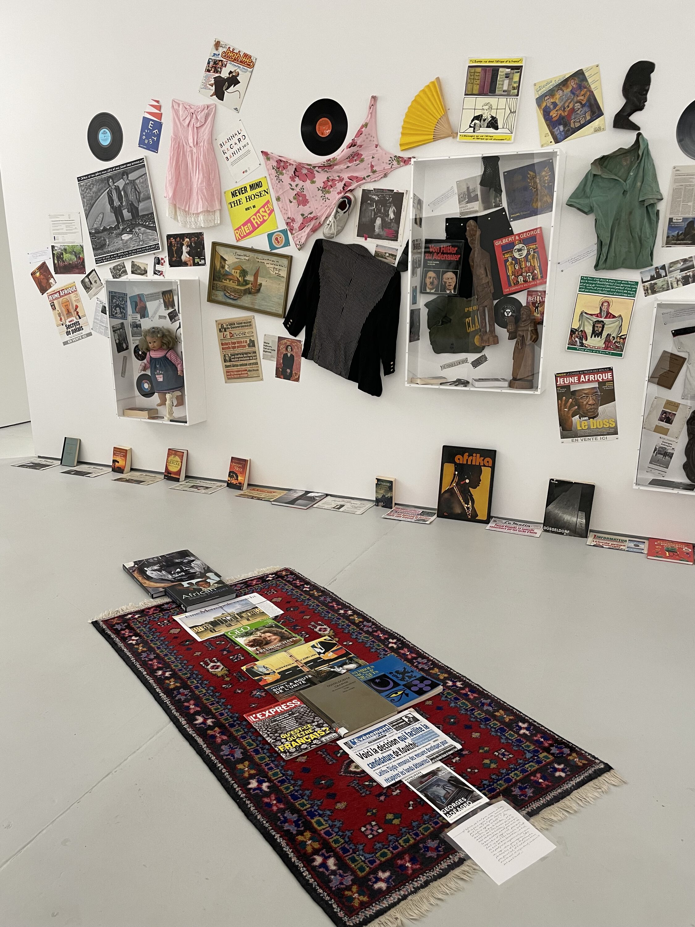 an installation on the floor, books newspapers on a carpet, paintings, textiles objects on the wall behind.