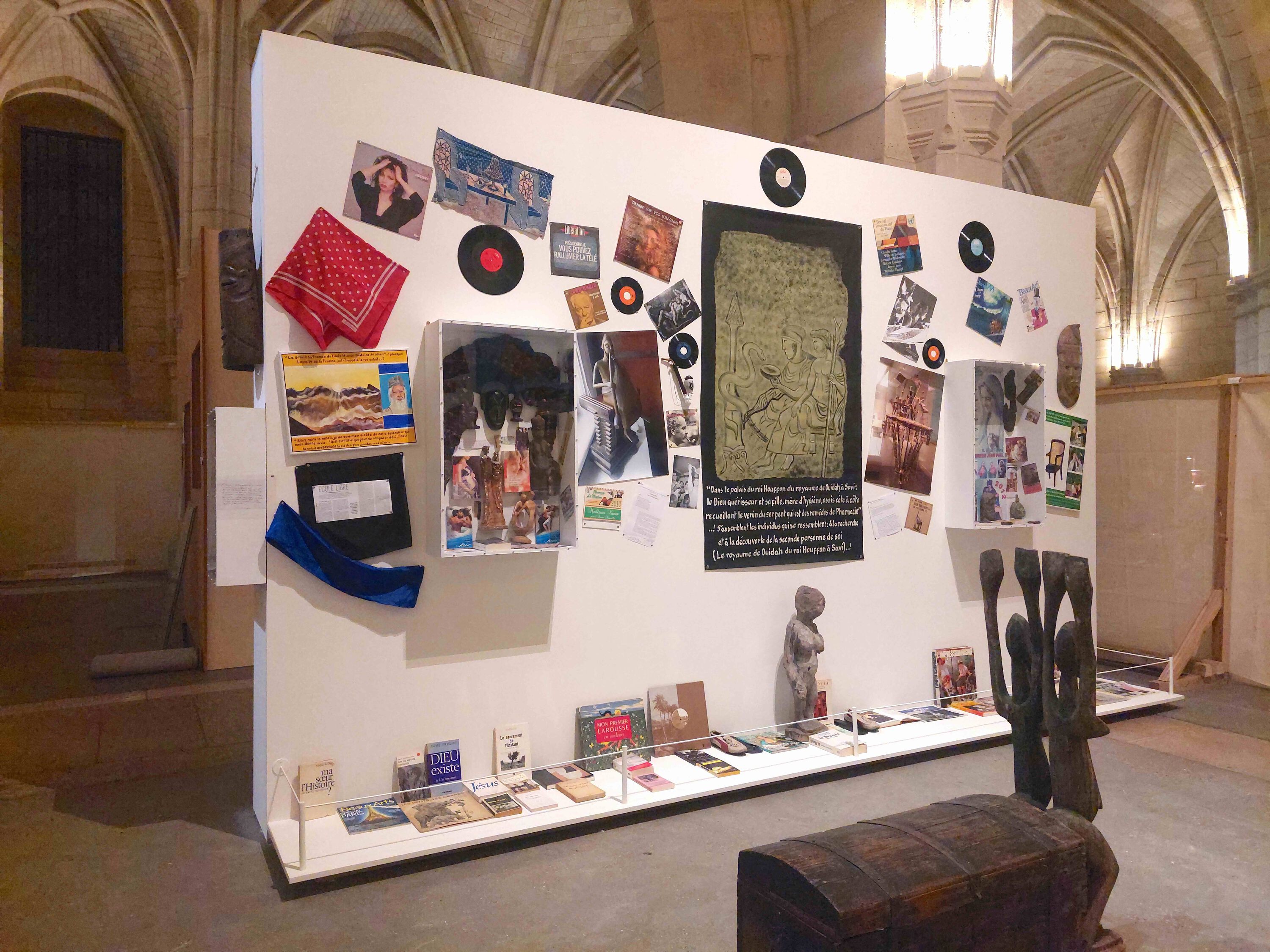 in a hall with gothic vaults, a white wall with numerous paintings photos and vinyl records.