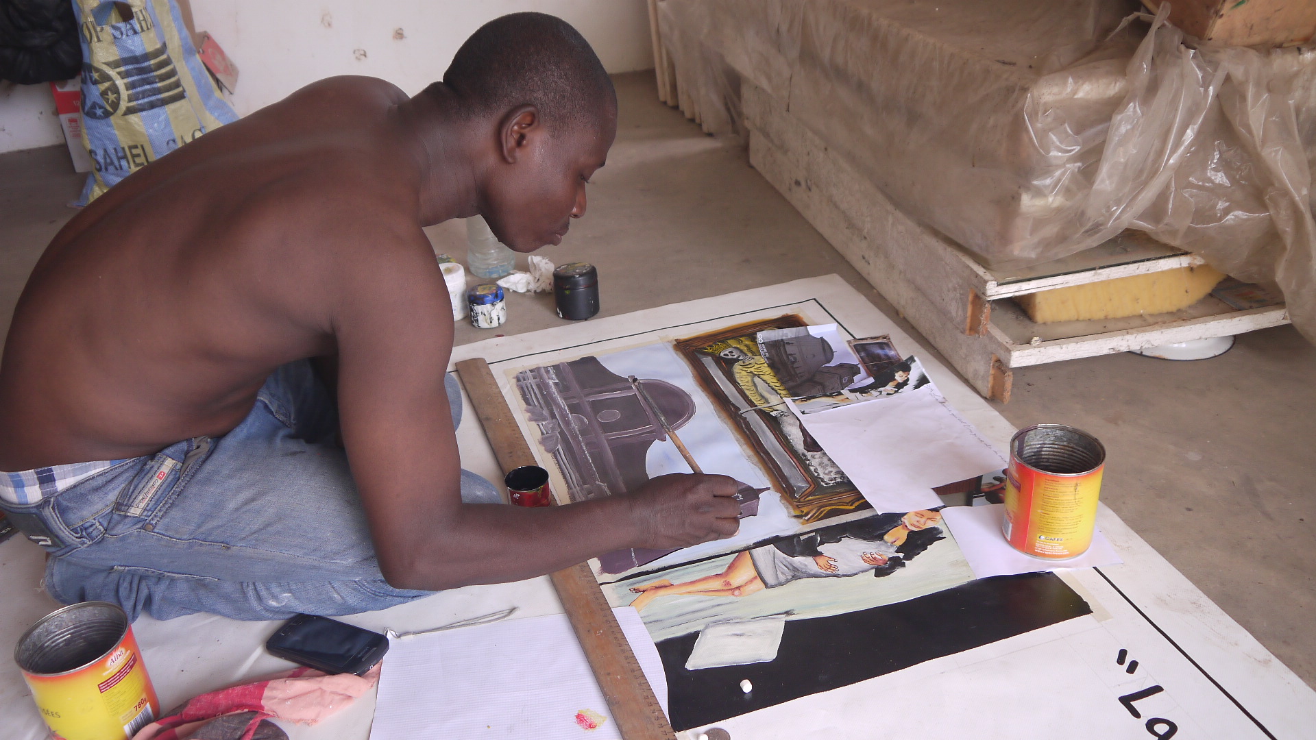 a painter kneels on the floor and copies images that Adéagbo gave him on a big vinyl banner