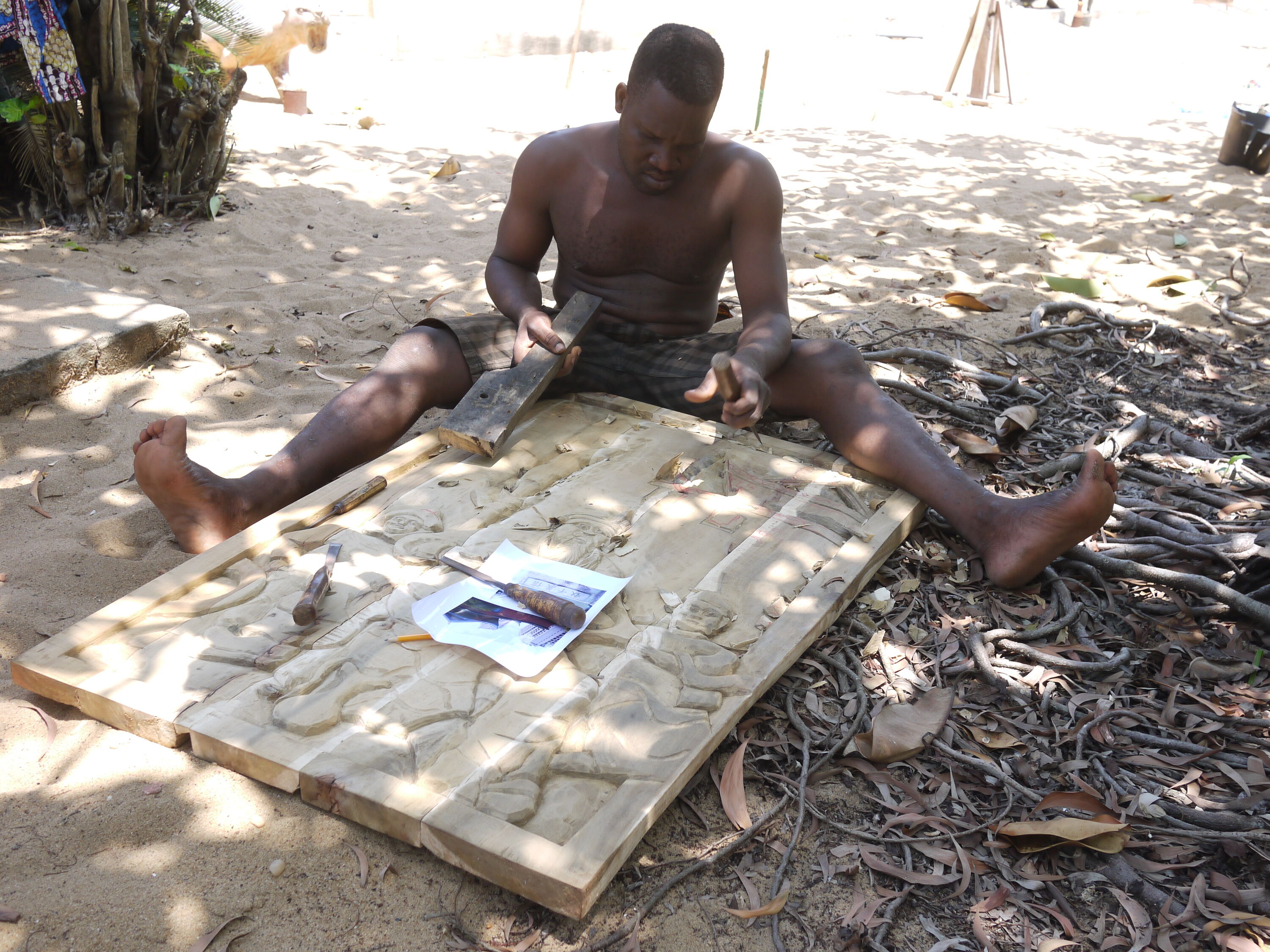 artisan Hugues Hountondji sits on floor and carves on a large wooden panel